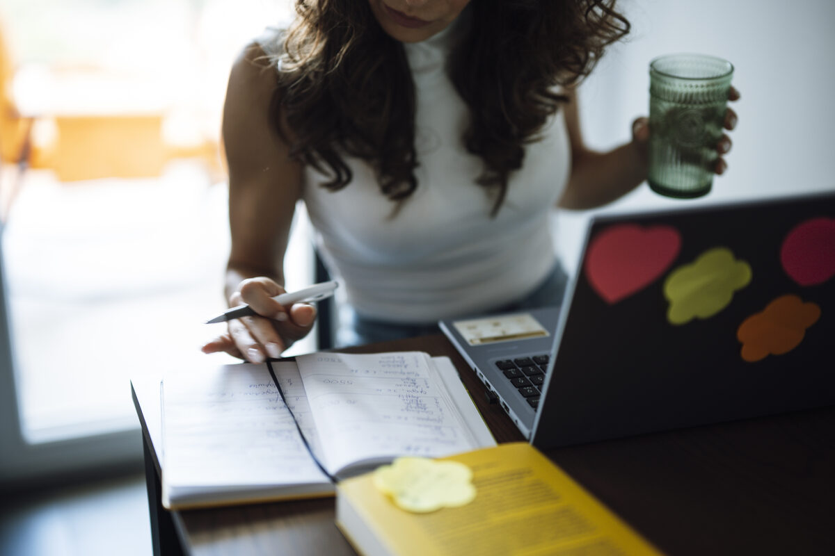 A woman writes in a physical notebook next to an open laptop on a desk, with a book and sticky note nearby, the kind of mixed paper-and-digital notes a workspace tool like Notion is meant to organize